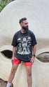 Man wearing a graphic t-shirt and red shorts standing in front of a large concrete structure.
